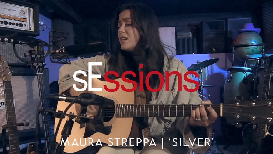 Maura Streppa in a recording room playing a guitar with drums, speakers, and guitars, a globe and cowboy hat in the background. There is a purple hue in the room