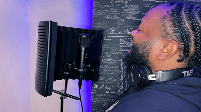 Studio setting with singer performing with a smile on his face. He is singing into an sE2200 with a cardioid polar pattern and RF PRO Reflexion Filter with a bluish purple light and sound proofing with a written sketch design.