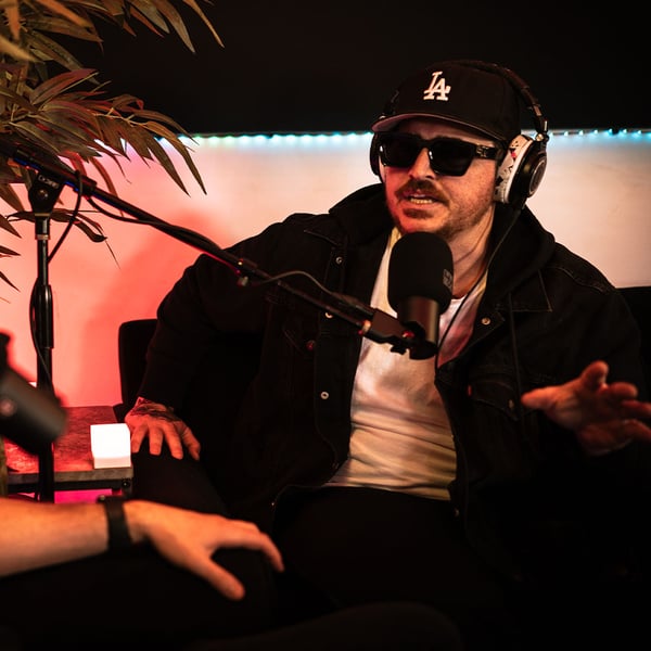 male podcaster wearing a black LA hat, sunglasses, headphones with white fluffy ear pads, white shirt and black jacket. A DynaCaster mic with a black windscreen pointed at the podcaster. Green fern in the room with pinkish, orange lighting.