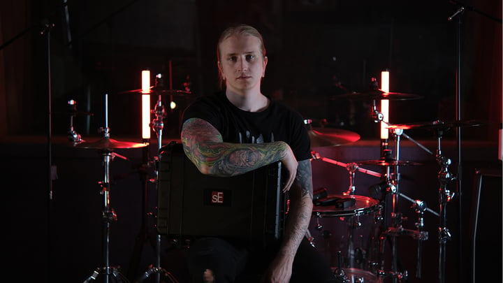 Drummer Maxim in his studio, sitting on a drum throne and holding the V CASE for drum mics, black molded touring case with a clear acrylic drum set in the background and red highlights.