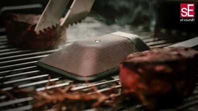 The BL8 on a BBQ pit surrounded by steaks with tongs in the image.