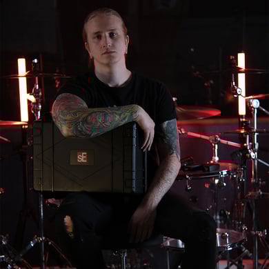 Drummer Maxim in his studio, sitting on a drum throne and holding the V CASE for drum mics, black molded touring case with a clear acrylic drum set in the background and red highlights.